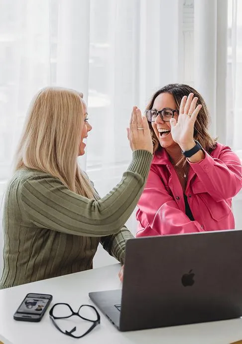 Jess Green and Natasha Roberson sharing a celebratory high five while working on a laptop in a bright, modern workspace with smiles and energy