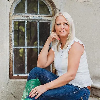Jessica Green sitting casually by an old window, wearing jeans and boots, smiling with a thoughtful pose.