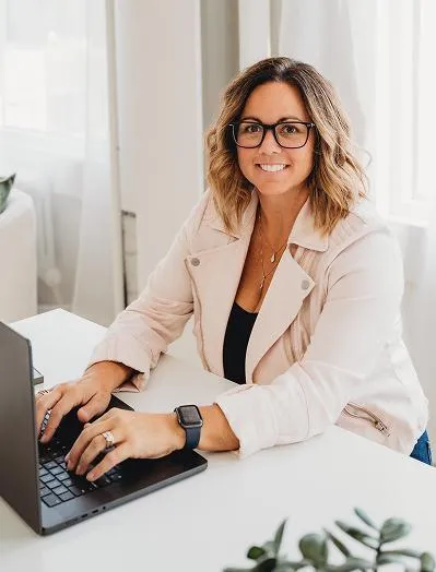 Natasha Roberson working at a laptop in a bright white office, wearing a light pink jacket and glasses