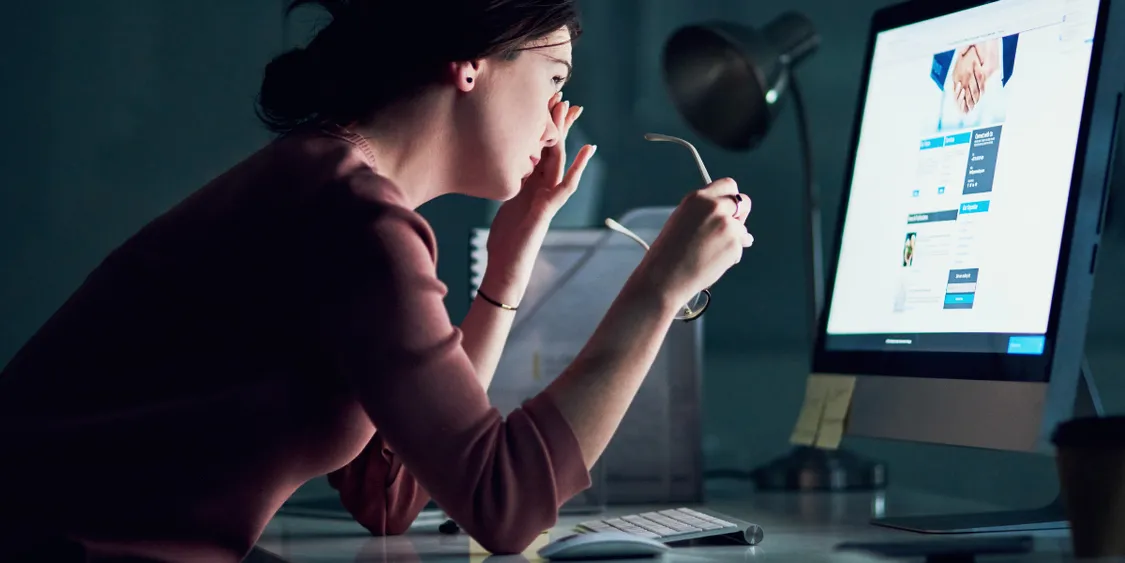 Woman sitting at a desk in a dark room, removing her glasses and rubbing her eyes while working late on a computer.