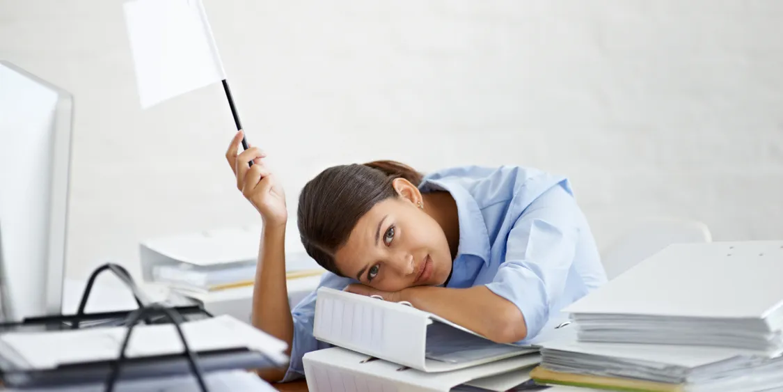 Tired woman resting her head on a stack of binders at her desk, holding a white flag in surrender.