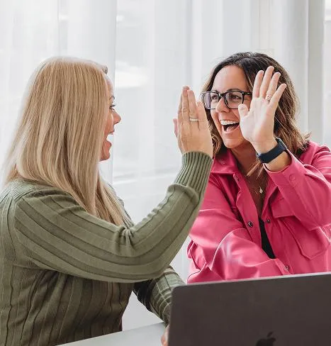 Jess Green and Natasha Roberson celebrating a win with a high five while sitting at a laptop, full of energy and collaboration in a modern workspace
