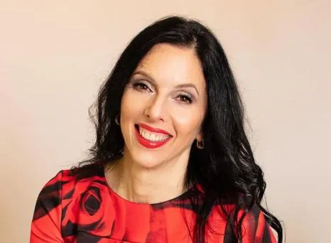 Woman with long dark hair and red lipstick smiling warmly in a floral red dress against a neutral background.
