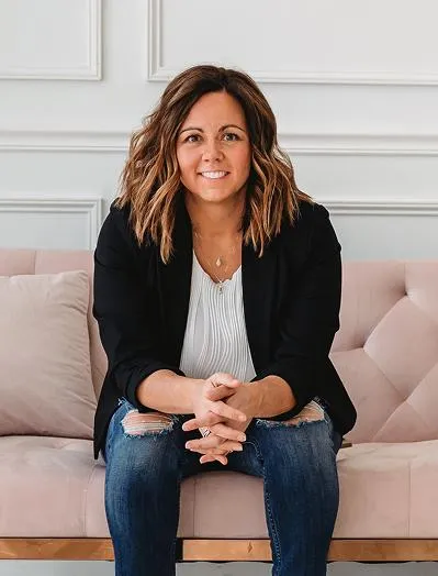 Natasha Roberson sitting on a pink couch in a black blazer and jeans, smiling with hands clasped