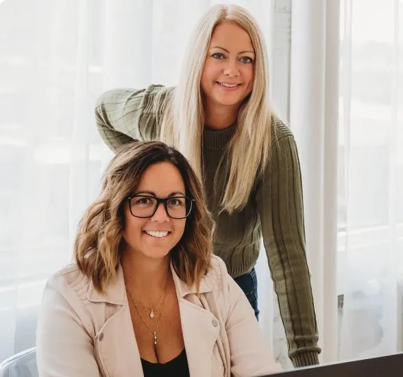 Jess Green and Natasha Roberson smiling together at a bright office desk, with Natasha seated in front of a laptop and Jess standing beside her in a green sweater