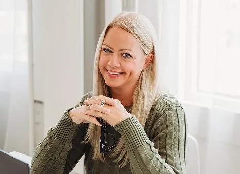 Professional woman with long blonde hair smiling at her desk, hands folded and confident expression.
