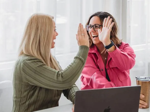Two women smiling and high-fiving at a desk with a laptop, celebrating a business win together.