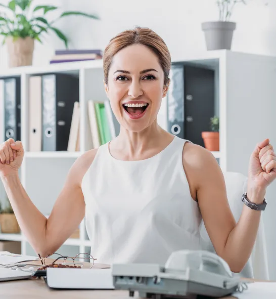 xcited professional woman celebrating success at her desk, wearing a white sleeveless top in a bright office environment with binders, books, and a telephone on her desk.