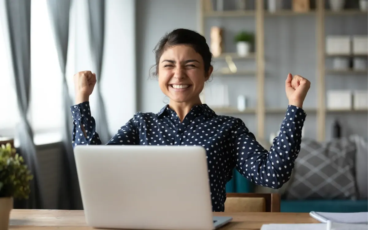 Smiling woman raising her fists in celebration while sitting at a desk with a laptop