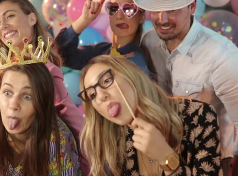  lively group of partygoers strikes silly poses with fun props, including crowns, glasses, and hats, in front of a festive balloon backdrop. Perfect for showcasing interactive photo booth experiences for weddings, birthdays, and corporate events in College Station, Austin, Dallas, Houston and San Antonio.