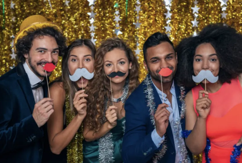 A well-dressed group of five people holding mustache and lips props in front of a glamorous gold backdrop, striking playful poses. Perfect for weddings, corporate parties, and high-end events in Texas.