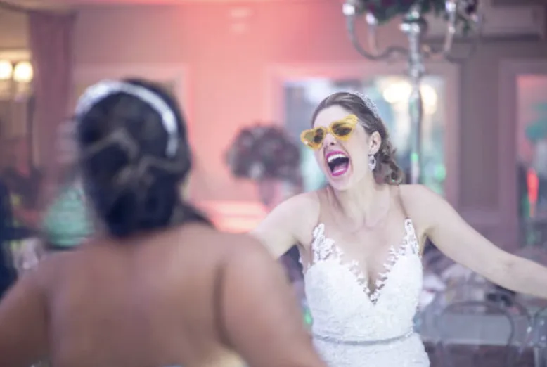 A bride in heart-shaped sunglasses joyfully dances and laughs, embracing the fun and excitement of a DJ-driven wedding party.