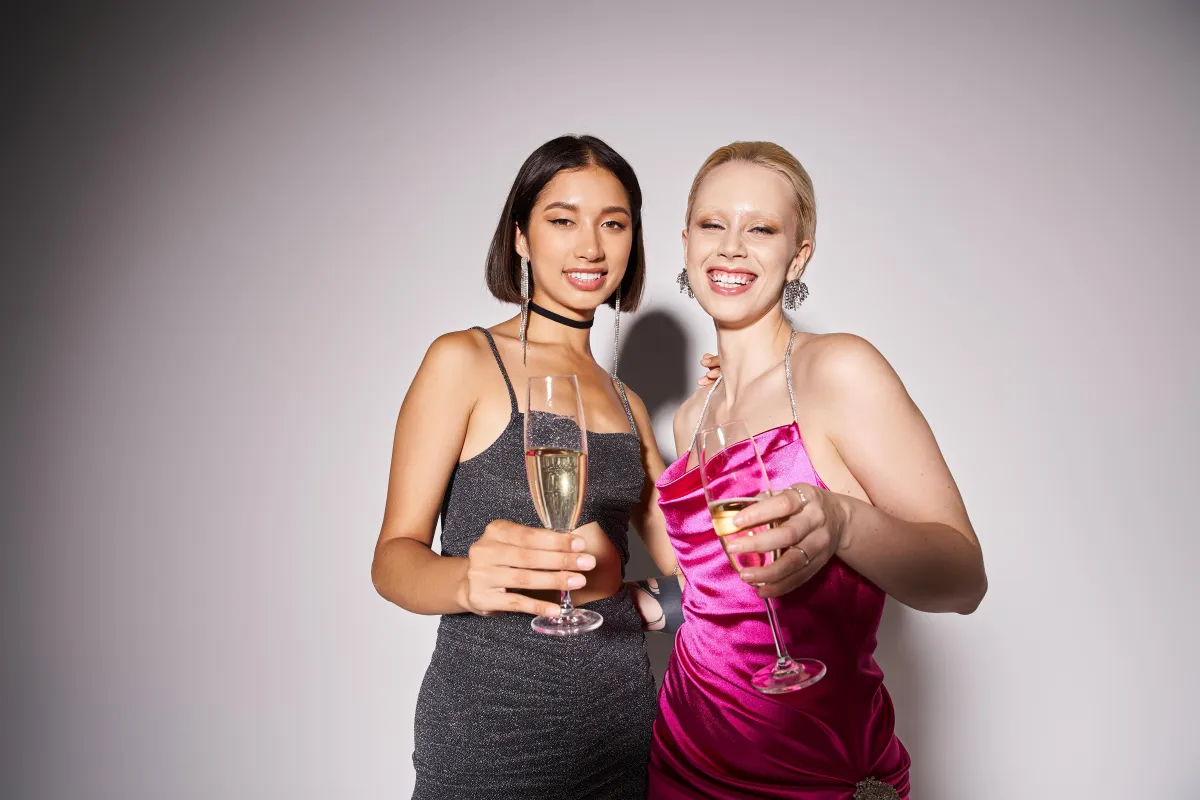  Two fashionable women in cocktail dresses holding champagne glasses and posing in a modern photo booth with a sleek, minimal backdrop, perfect for upscale events. In Glam Booth Style