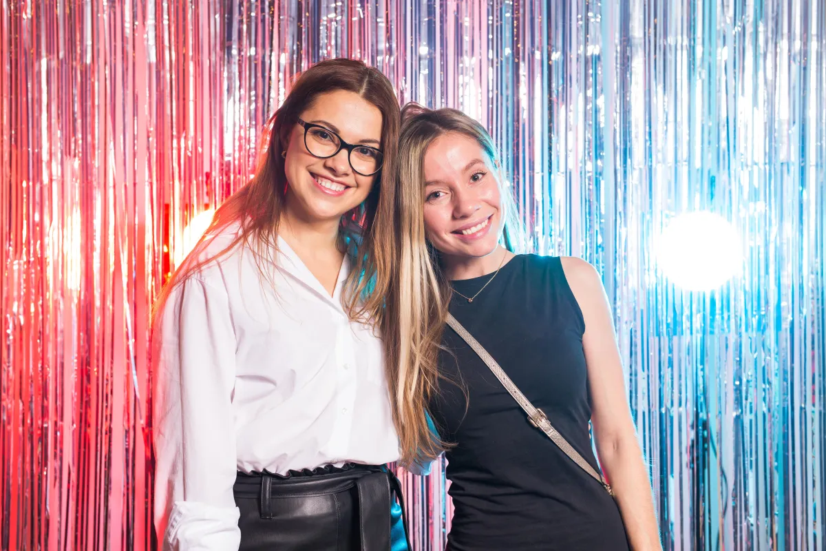 Two smiling women standing close together in front of a dazzling red, silver, and blue metallic tinsel backdrop, capturing a fun and stylish photo booth moment at an event in Texas.