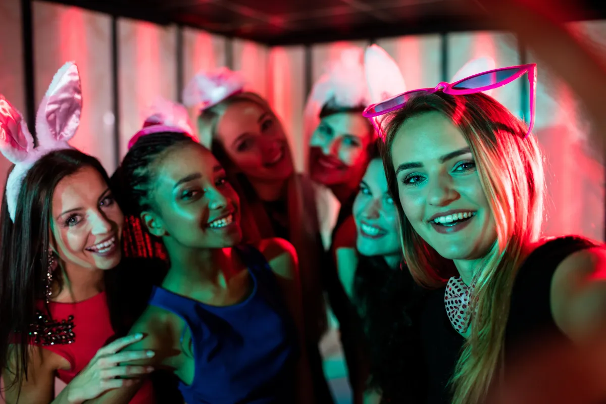 Group of smiling women at a bachelorette party taking a selfie, wearing bunny ears and fun accessories under colorful nightclub lighting.