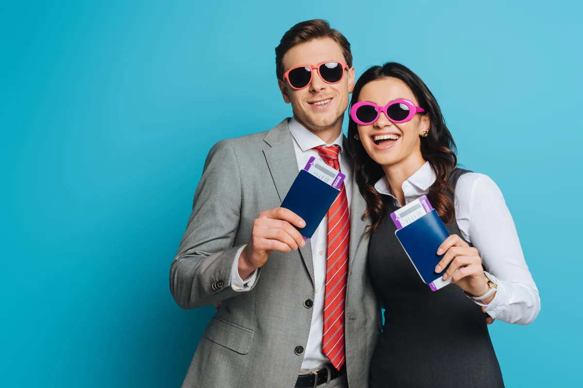 Smiling couple in business attire wearing fun sunglasses, holding passports and boarding passes, ready for a honeymoon or destination event.