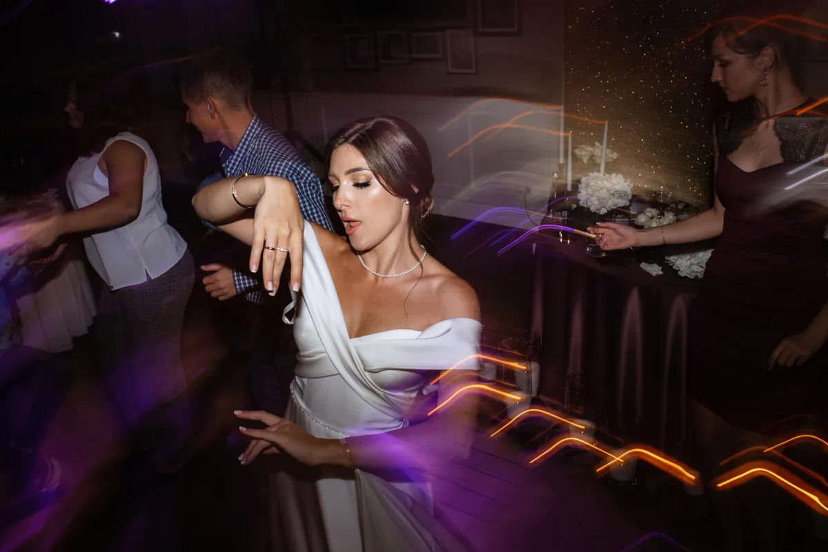 A radiant bride dances with joy at her wedding reception, surrounded by guests and dynamic DJ lighting effects that set the perfect party atmosphere.