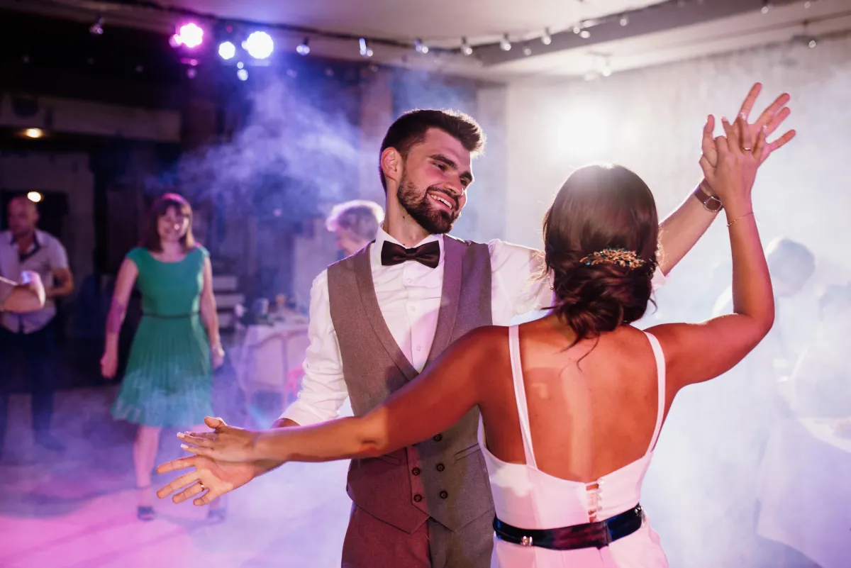 A newlywed couple enjoys their first dance in a dreamy setting, enhanced by soft smoke and colorful DJ lighting.