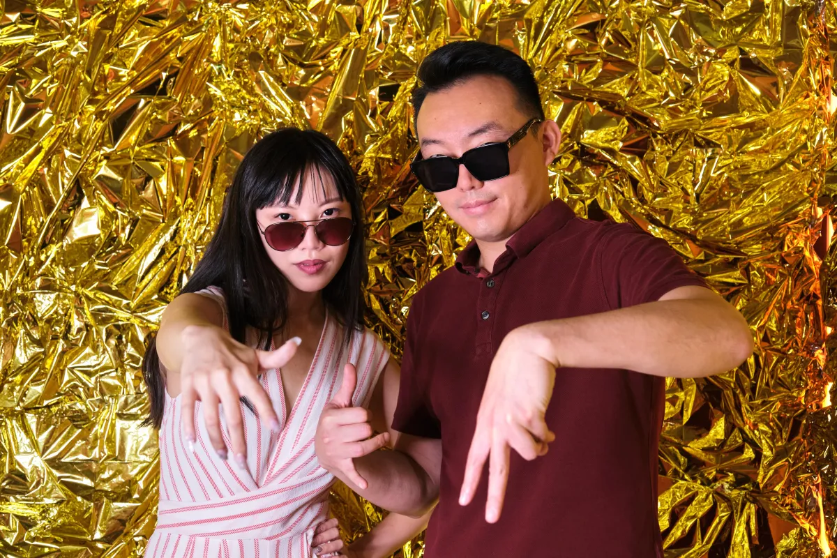 Two stylish guests wearing sunglasses posing in front of a gold metallic photo booth backdrop at an event.