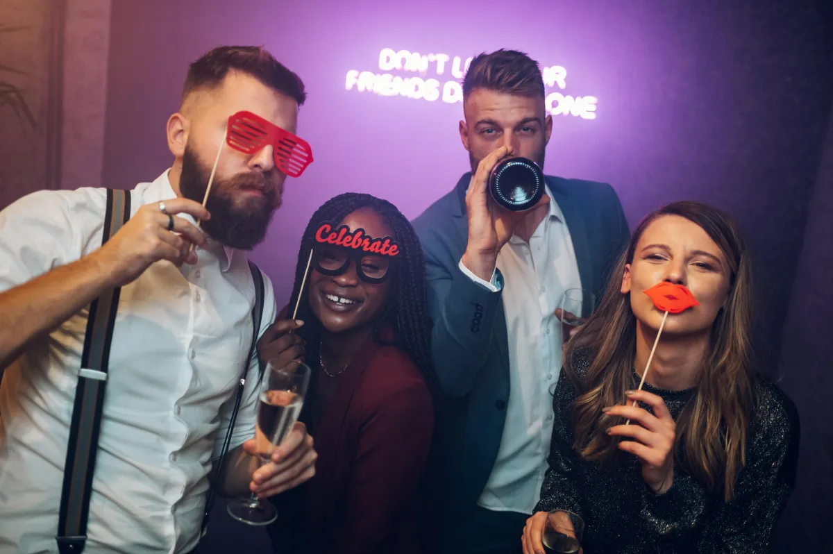  Four well-dressed friends at a luxury event using fun photo booth props, including oversized glasses and a champagne toast, with a neon "Don’t Let Your Friends Dance Alone" sign in the background. Ideal for high-end weddings and corporate events in Texas.