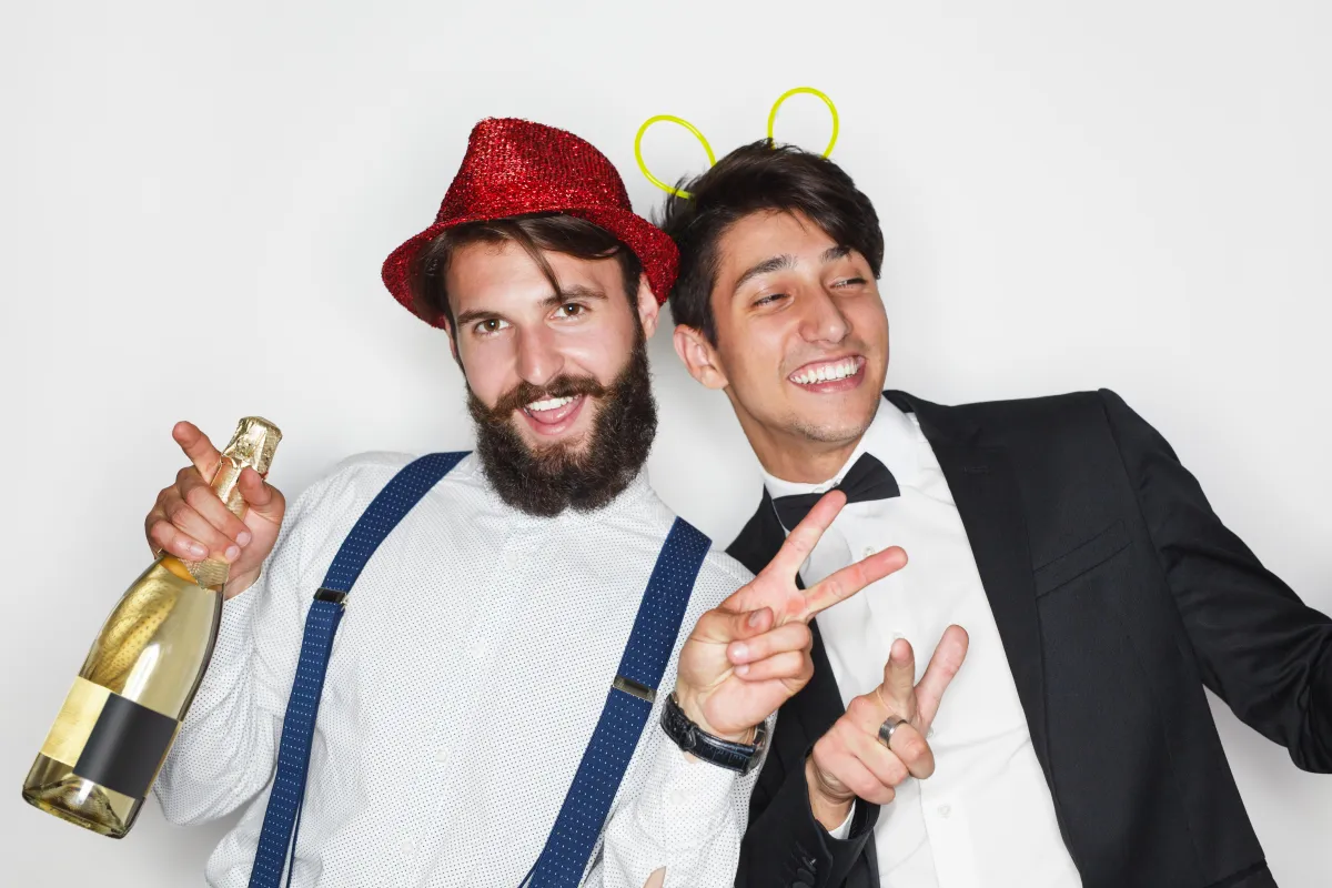 Two men celebrating at a wedding photo booth in San Antonio, Texas, dressed in fun party attire with champagne and props.