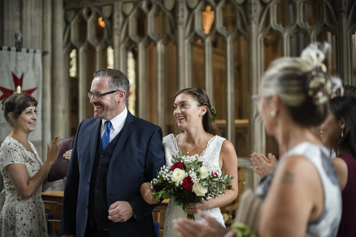 A joyful bride walks arm-in-arm with an older gentleman, possibly her father, down the aisle of a stunning gothic-style church after the wedding ceremony. Guests cheer and clap as she holds a beautiful bouquet of red and white roses, radiating happiness.