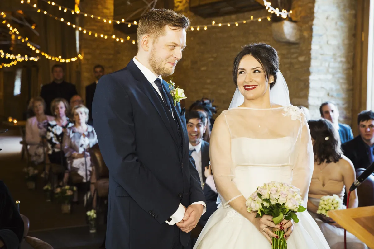 A newlywed couple shares a joyful moment during their rustic wedding ceremony in a warmly lit barn venue adorned with string lights. The groom, dressed in a navy suit, lovingly gazes at his radiant bride, who wears an elegant white gown with sheer detailing and holds a bouquet of soft pink roses. Guests in the background watch with smiles as the couple celebrates their special day.