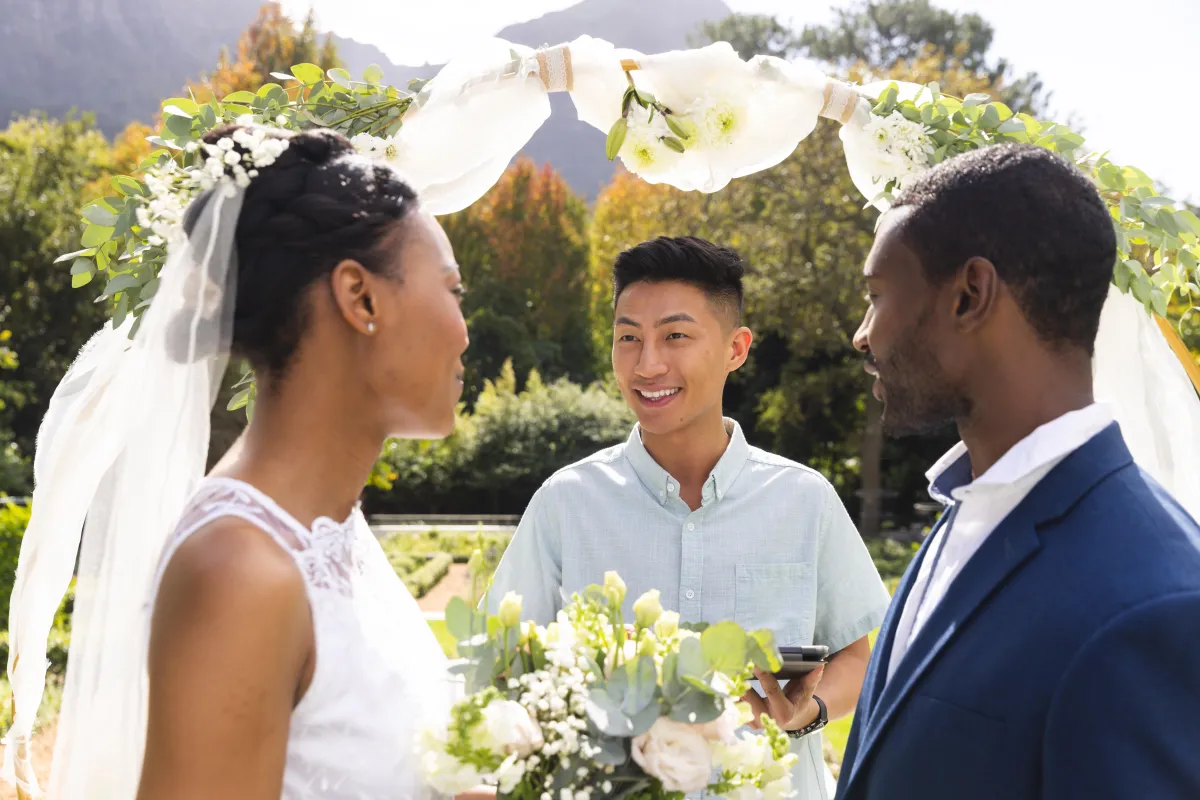 A happy bride and groom stand under a beautifully decorated floral wedding arch, exchanging smiles with their officiant during an intimate outdoor ceremony with lush greenery and mountains in the background.