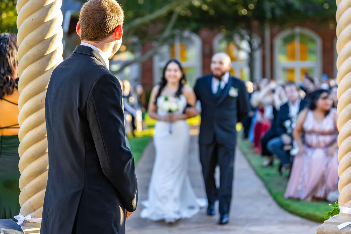 Groom watching his bride walk down the aisle with her father at an outdoor wedding ceremony, surrounded by guests.