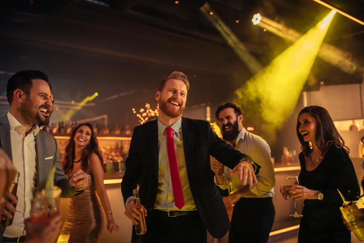 A lively group of stylish professionals laughing and dancing at an upscale nightclub, illuminated by warm golden lighting with a vibrant party atmosphere.