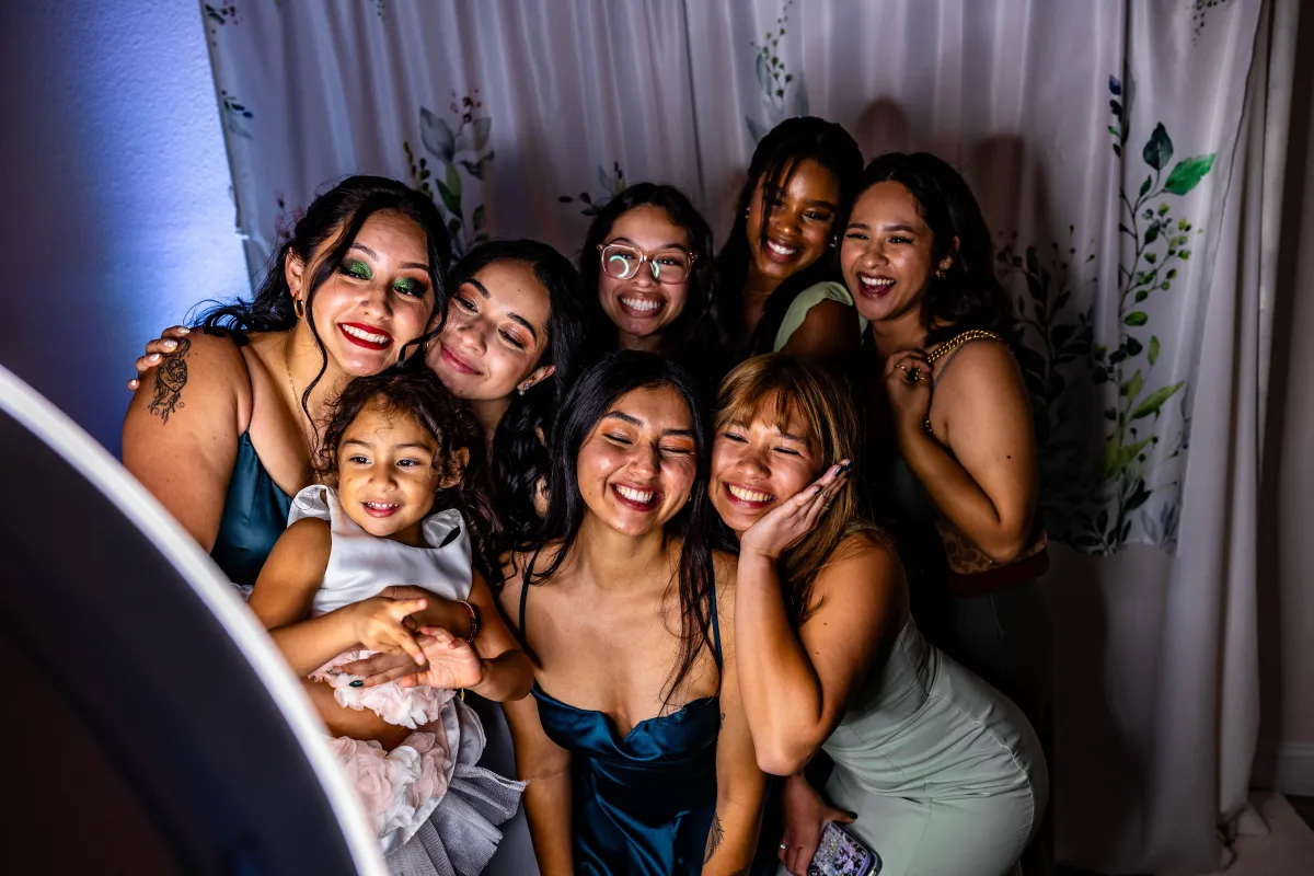 Group of smiling women and a child posing for a fun photo booth session at an event with a floral backdrop.