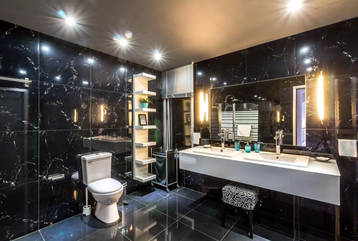 Luxuriöses Badezimmer mit schwarzen Marmorwänden und glänzenden Bodenfliesen. Die moderne Ausstattung umfasst ein Doppelwaschbecken mit großem Spiegel und integrierter Beleuchtung, ein wandmontiertes Regal mit Dekoration und eine Toilette. Die stimmungsvolle Beleuchtung und eleganten Materialien verleihen dem Raum eine edle, zeitgenössische Atmosphäre.