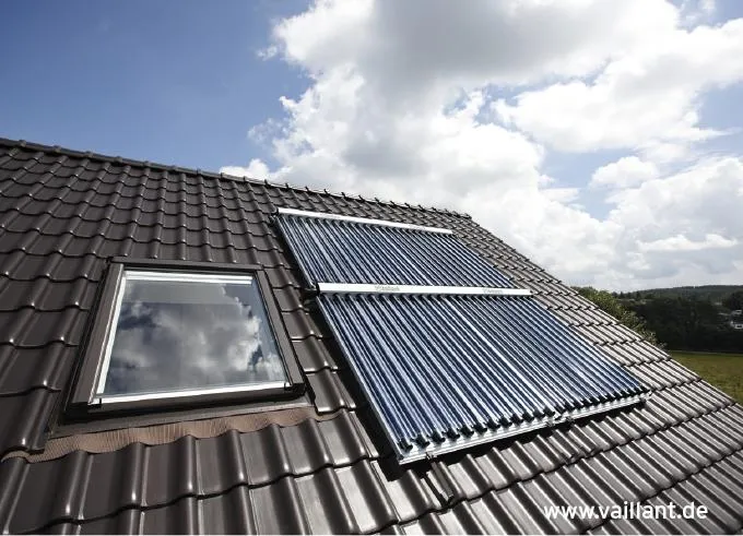 Vaillant Solarkollektoren auf einem mit dunklen Ziegeln gedeckten Schrägdach neben einem Dachfenster. Die Vakuumröhrenkollektoren nutzen Sonnenenergie zur umweltfreundlichen Wärmegewinnung. Der Himmel mit Wolken spiegelt sich im Fenster und sorgt für ein harmonisches Bild von moderner Solarenergie und traditioneller Architektur.