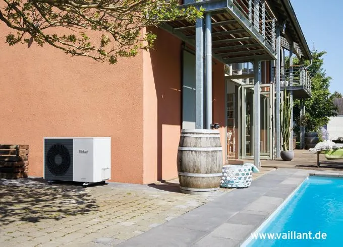 Vaillant Luft-Wasser-Wärmepumpe installiert im Außenbereich eines modernen Hauses mit oranger Fassade, nahe einem Pool. Die Wärmepumpe steht auf einer gepflasterten Terrasse neben einem dekorativen Holzfass. Die Umgebung umfasst eine moderne Architektur mit großen Glasflächen und einem angrenzenden Garten.