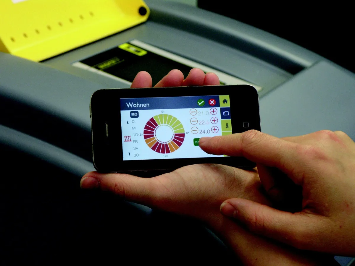 Smartphone mit App zur Steuerung der Heizungsanlage. Eine Hand bedient die Benutzeroberfläche, die eine Heizkurve anzeigt, mit der sich die Raumtemperaturen anpassen lassen. Die Steuerung erfolgt per Fingertipp, um die gewünschte Temperatur in verschiedenen Zeiträumen festzulegen. Im Hintergrund ist die Heizungsanlage zu sehen.