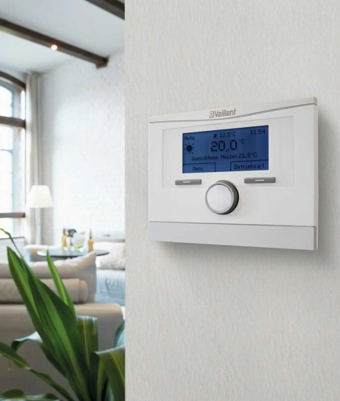 Moderne Heizungssteuerung von Vaillant an einer weißen Wand in einem hellen Wohnzimmer. Der digitale Thermostat zeigt eine Temperatur von 20°C an und bietet verschiedene Einstellungen zur individuellen Regelung der Raumtemperatur. Im Hintergrund sind ein gemütliches Sofa, eine Zimmerpflanze und große Fenster zu sehen, die das Zimmer mit natürlichem Licht durchfluten.