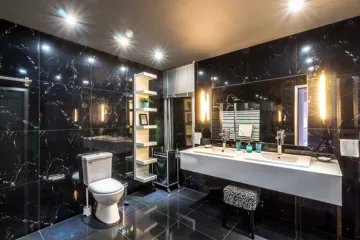 Luxuriöses Badezimmer mit schwarzen Marmorwänden und glänzenden Bodenfliesen. Die moderne Ausstattung umfasst ein Doppelwaschbecken mit großem Spiegel und integrierter Beleuchtung, ein wandmontiertes Regal mit Dekoration und eine Toilette. Die stimmungsvolle Beleuchtung und eleganten Materialien verleihen dem Raum eine edle, zeitgenössische Atmosphäre.