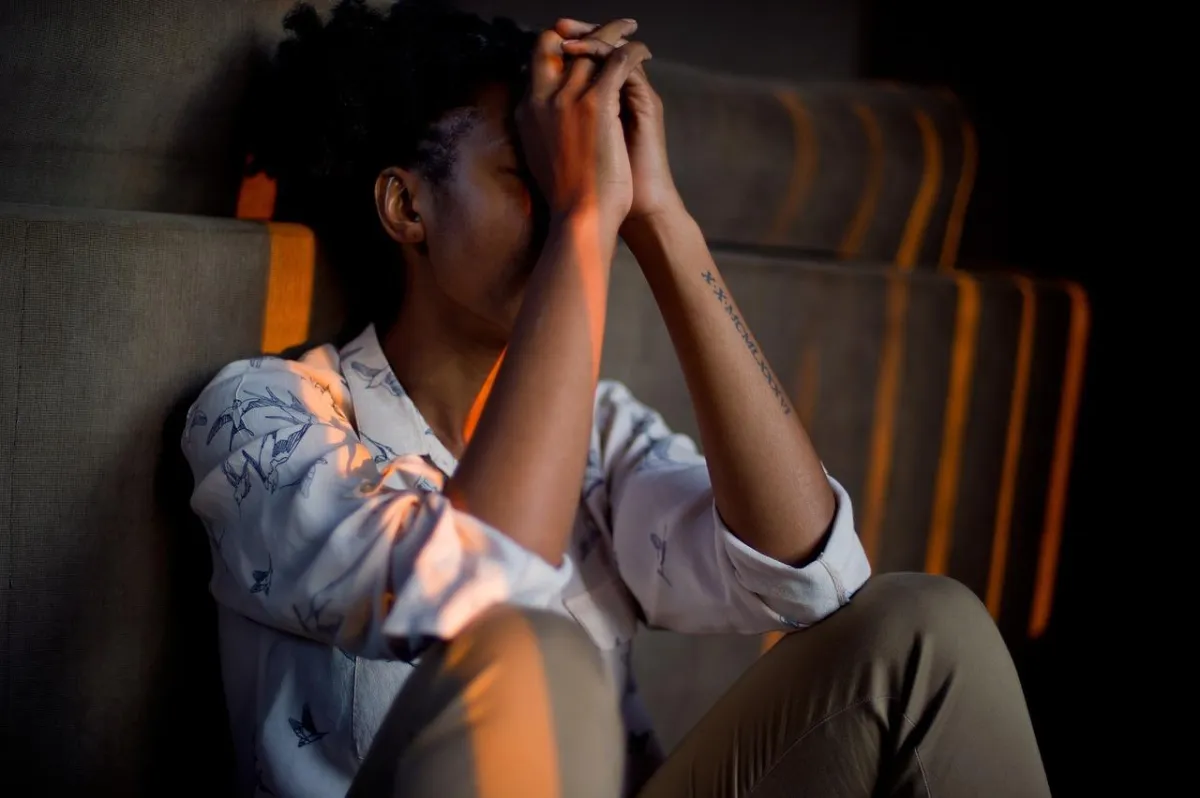 A person sits with hands over their face in a dimly lit room, wearing a bird-patterned shirt and brown pants. A tattoo with Roman numerals is on their forearm, and warm orange light casts shadows from the background slats.