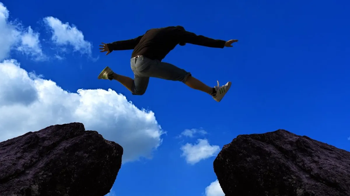 A person in mid-air jumps between two large rocks under a bright blue sky with scattered clouds. They wear a black long-sleeve shirt, denim shorts, and white sneakers with green stripes.
