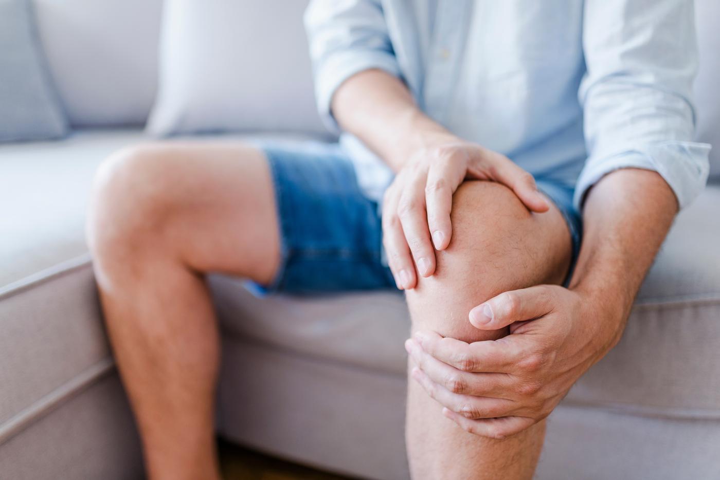 Close-up of a person's hands tightly grasping their right knee, wearing blue jeans, conveying knee pain