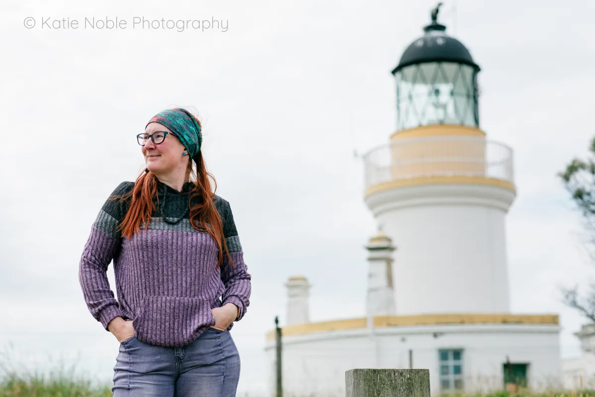 Photograph of a woman looking to the left with a lighthouse in the background. Photograph (C) Kaite Noble Photography