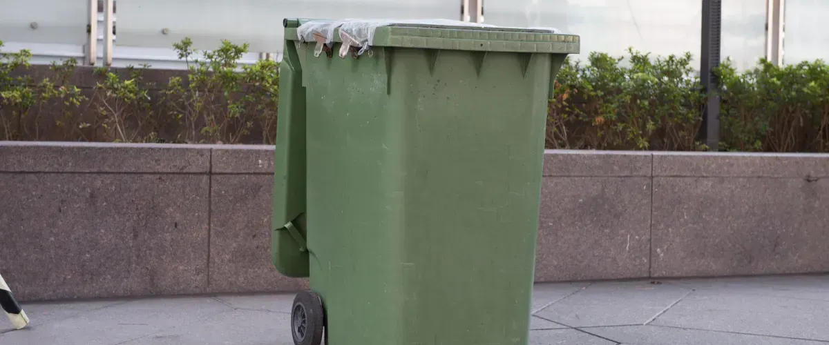 Dirty green trash bin on a sidewalk near an office building with plastic liner peeking from the lid.