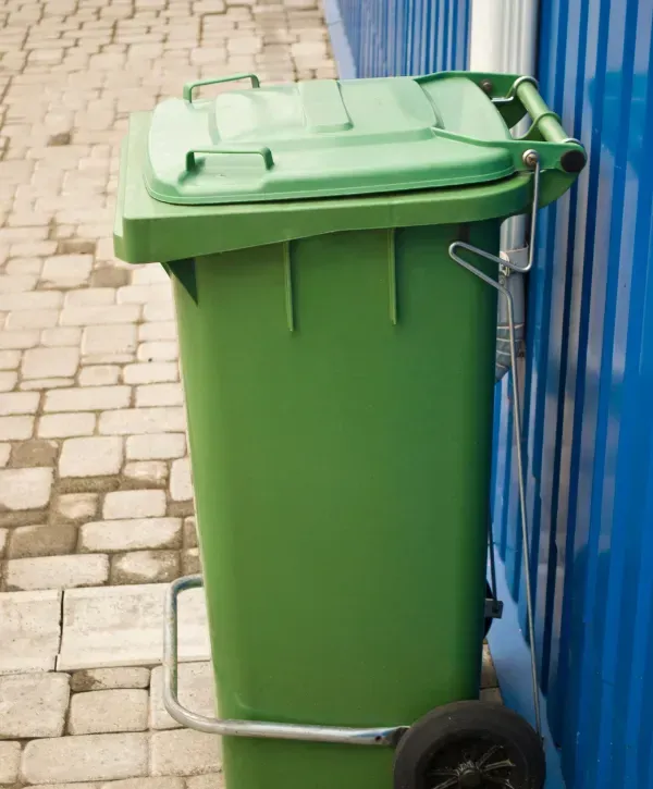 Single green trash bin with wheels and metal handle next to a blue building exterior.
