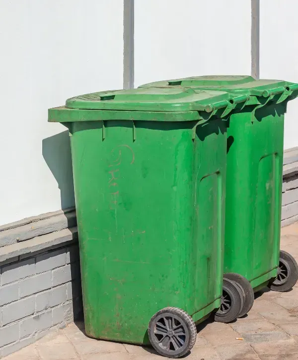 Two green trash bins placed on a brick sidewalk against a white wall.