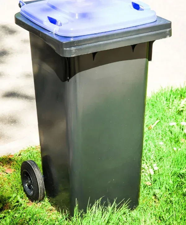 A dark green recycling bin with a light blue lid and a single wheel visible, sitting on green grass next to pavement.