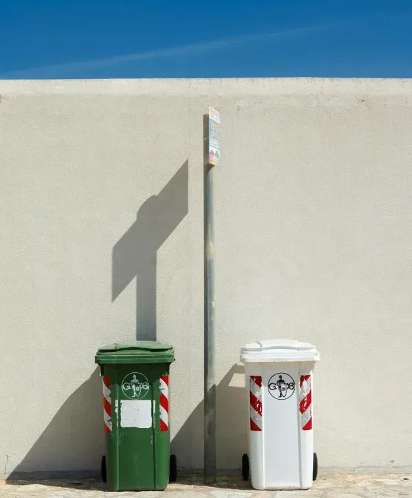 A green and a white refuse bin with red and white striped markings against a light wall and blue sky.