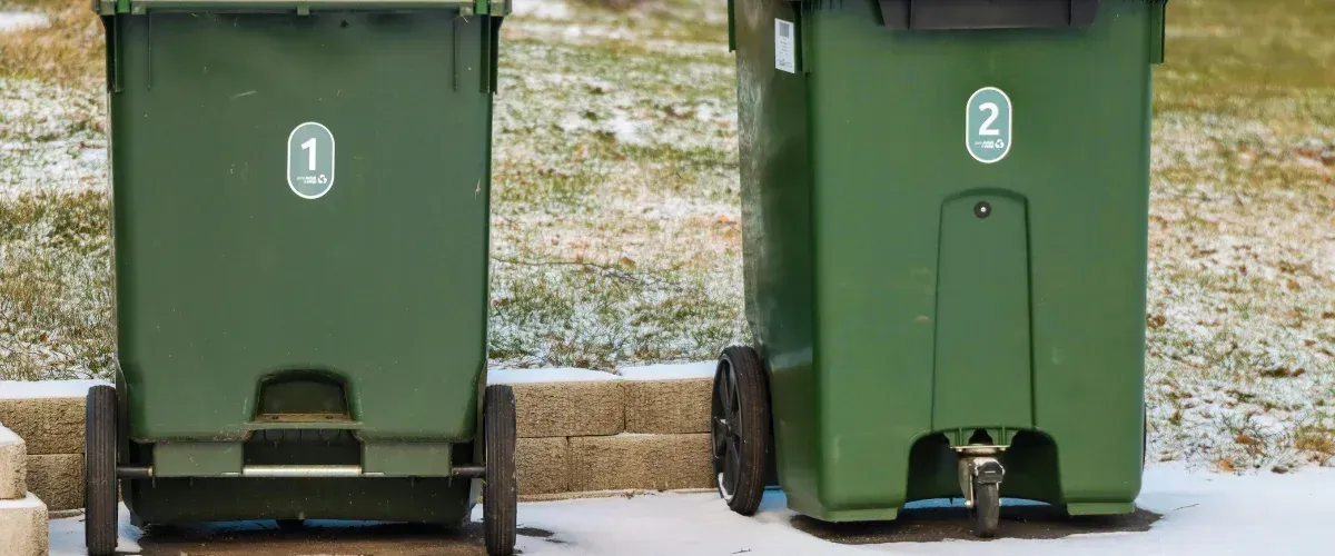 Two dark green recycling bins with numbers 1 and 2 on the front are standing on a snowy patch of grass.