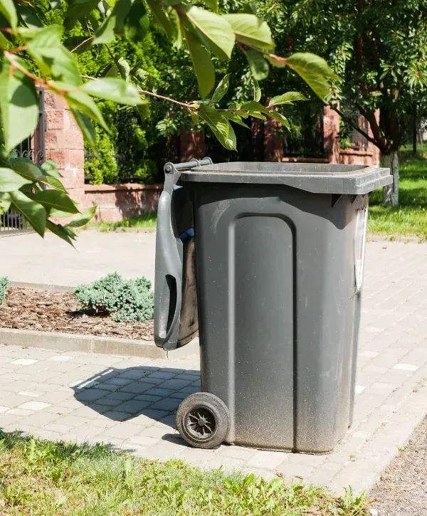 A dark gray plastic wheeled waste bin with its lid slightly ajar sits on a paved sidewalk on a sunny day.