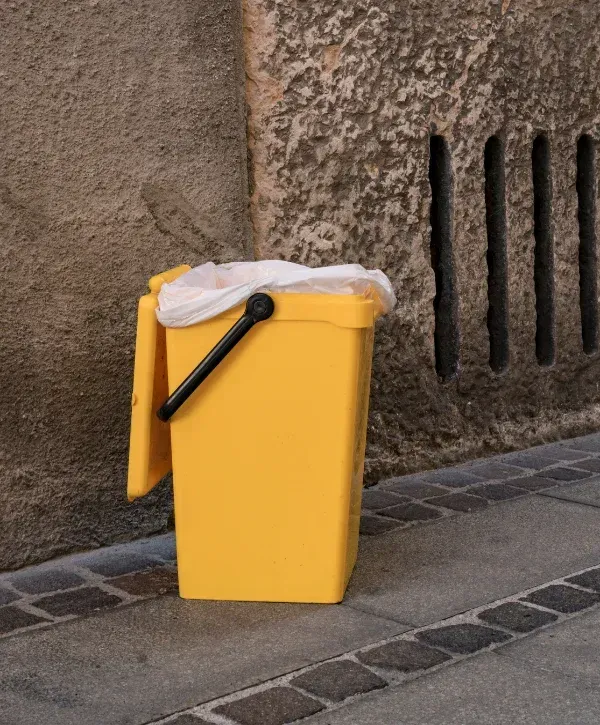 A bright yellow outdoor waste bin with a white plastic bag, set against a rough stone wall and cobblestone ground.