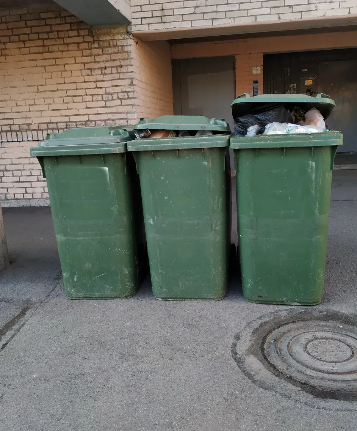 Three green dumpsters, two with closed lids and one overflowing with trash, stand against a brick building wall on asphalt.